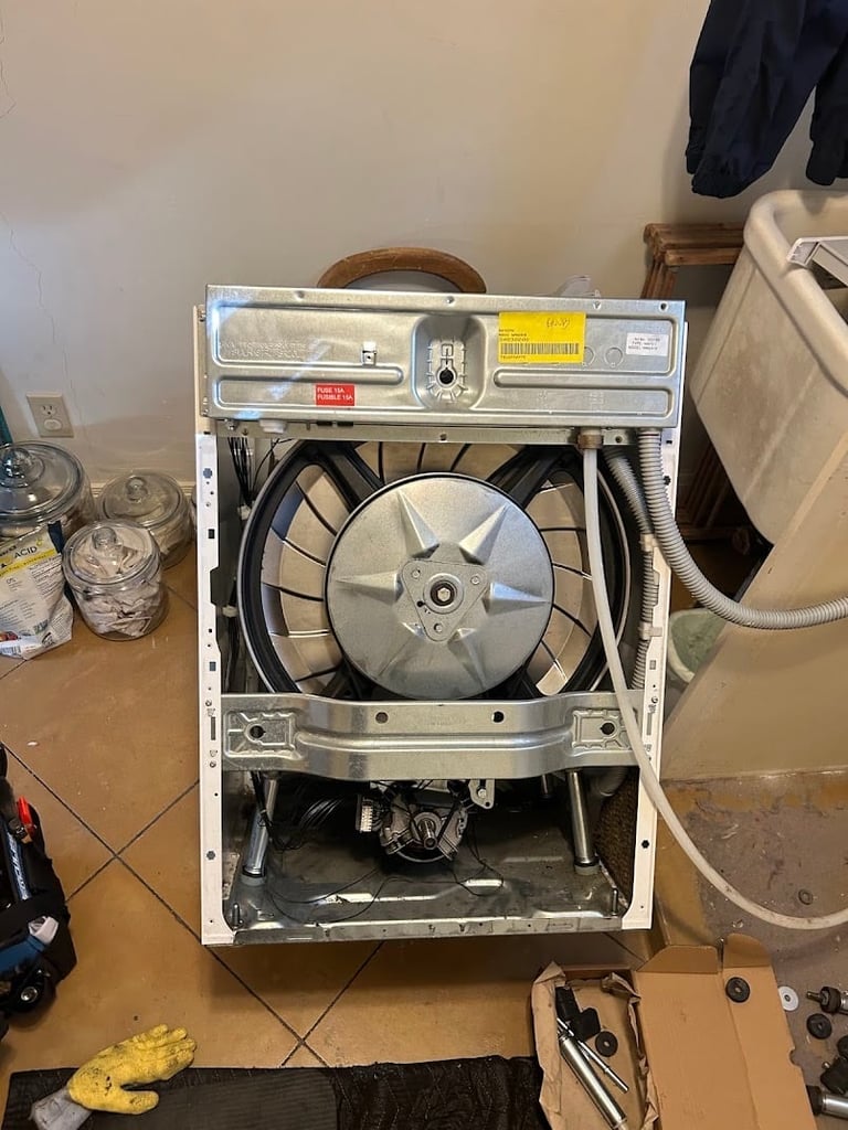 Open washing machine with exposed drum and internal components on wooden surface with laundry items around